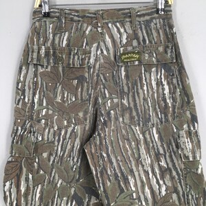 May include: Camouflage cargo pants with a Realtree Spartan label. The pants feature a woodland camouflage pattern in shades of green, brown, and white. The pants have multiple pockets and a button closure.