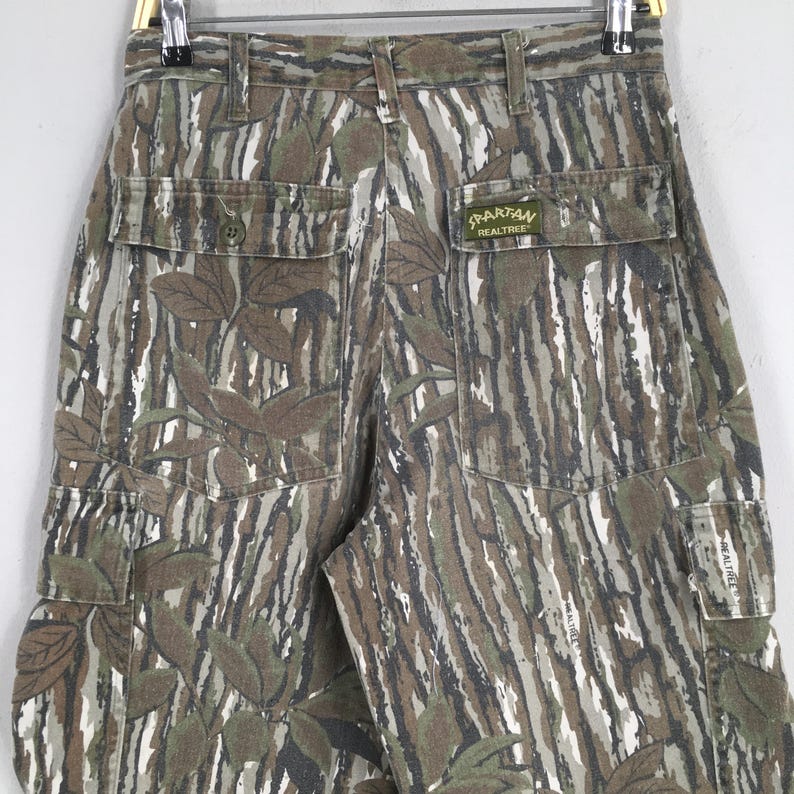 May include: Camouflage cargo pants with a Realtree Spartan label. The pants feature a woodland camouflage pattern in shades of green, brown, and white. The pants have multiple pockets and a button closure.