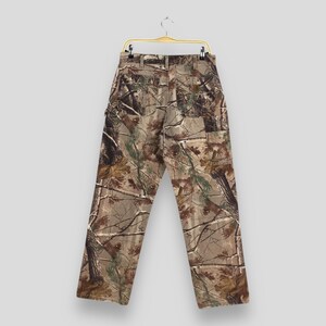 K&ouml;nnte beinhalten: Tarnhose, die an einem goldenen Kleiderb&uuml;gel h&auml;ngt. Die Hose zeigt einen realistischen Walddruck in Braun-, Gr&uuml;n- und Beiget&ouml;nen. Die R&uuml;ckseite der Hose ist sichtbar und zeigt Taschen und G&uuml;rtelschlaufen. Die Hose ist aus strapazierf&auml;higem Stoff.