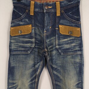 May include: Blue denim jeans with brown suede-like pocket accents and belt loops. The jeans have a faded wash and decorative stitching. The pockets have metal button closures. The waistband features a patterned fabric detail.