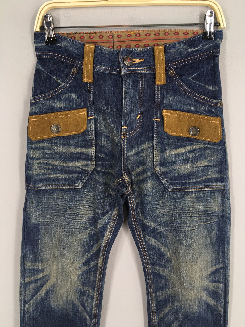 May include: Blue denim jeans with brown suede-like pocket accents and belt loops. The jeans have a faded wash and decorative stitching. The pockets have metal button closures. The waistband features a patterned fabric detail.