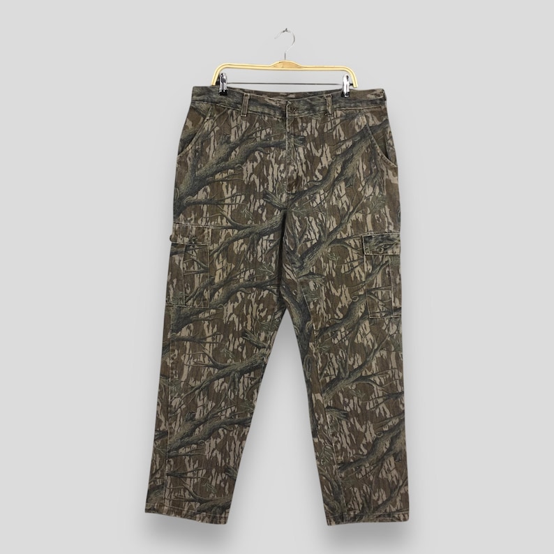 May include: Camouflage cargo pants with multiple pockets. The pants feature a brown, green, and gray tree branch pattern. The pants are hanging on a gold hanger.