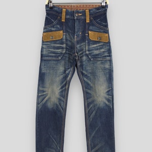 May include: Blue denim jeans with a faded wash and brown suede-like pocket details. The jeans have a classic five-pocket design with a button and zipper closure. The jeans are hanging on a gold hanger.