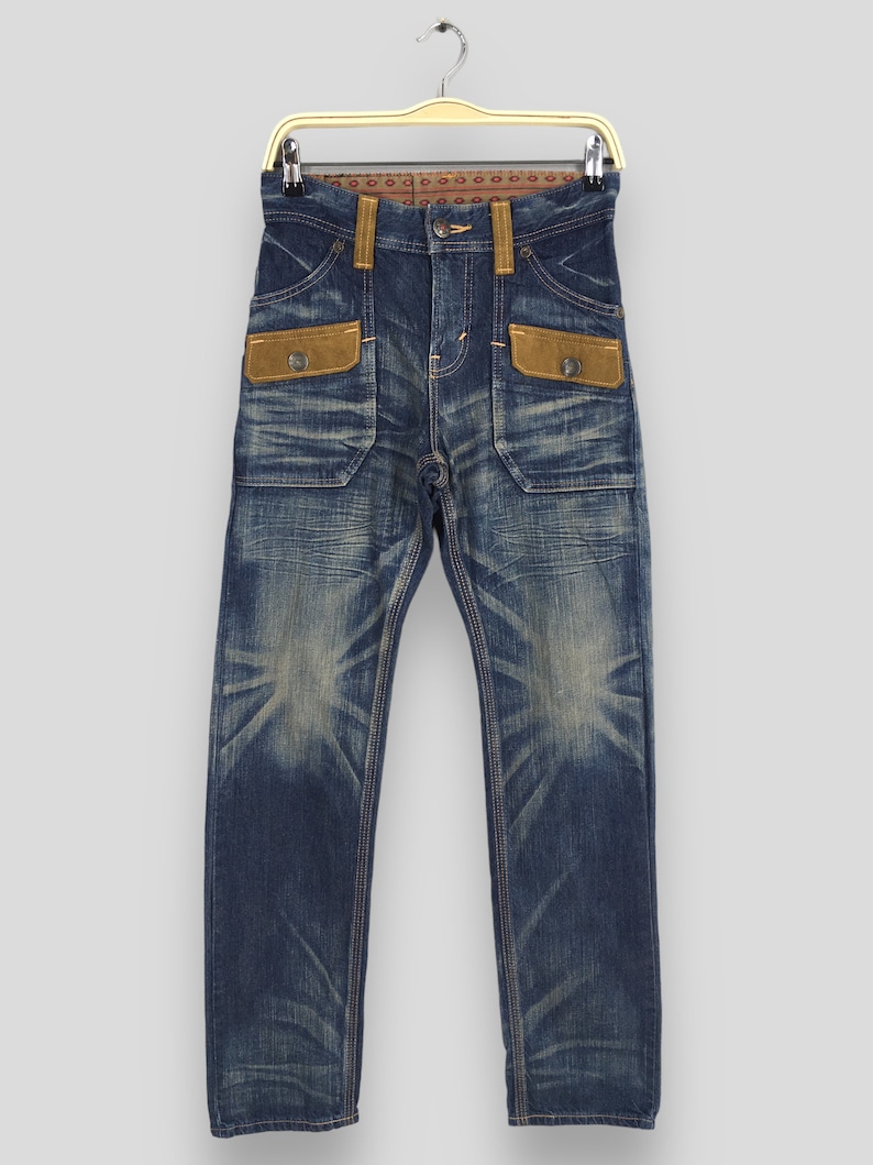 May include: Blue denim jeans with a faded wash and brown suede-like pocket details. The jeans have a classic five-pocket design with a button and zipper closure. The jeans are hanging on a gold hanger.