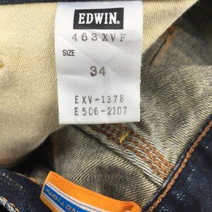 May include: Close-up of a dark blue denim jeans label with the brand name "EDWIN." The label displays the size "34" and other product information. An orange and blue tag with "XV-FUNCTION" is also visible.