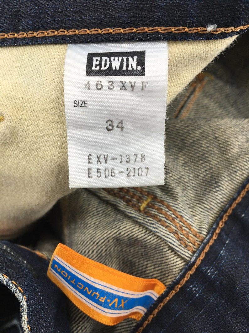 May include: Close-up of a dark blue denim jeans label with the brand name "EDWIN." The label displays the size "34" and other product information. An orange and blue tag with "XV-FUNCTION" is also visible.