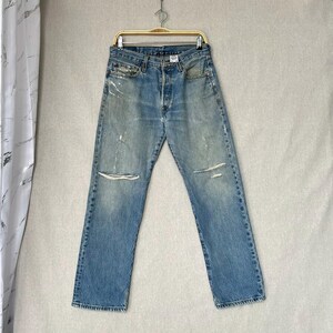 Könnte beinhalten: Hellblaue Jeans aus Denim mit Used-Details, einschließlich Knie-Rissen und verblichenen Flecken. Die Jeans hängen an einem goldfarbenen Kleiderbügel vor einem neutralen Hintergrund. Sie haben einen Knopfverschluss und ein klassisches Fünf-Taschen-Design.