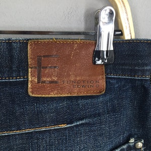 Puede incluir: Primer plano de unos vaqueros de mezclilla azul oscuro con un parche de cuero marr&oacute;n. El parche presenta el nombre de la marca "FUNCTION EDWIN" y un logotipo "F" estilizado. Los vaqueros est&aacute;n colgados de una percha de madera.