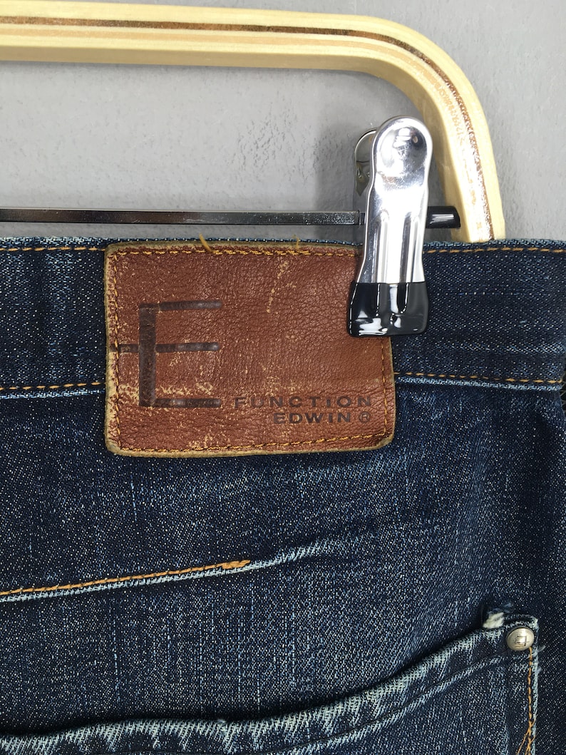 Puede incluir: Primer plano de unos vaqueros de mezclilla azul oscuro con un parche de cuero marr&oacute;n. El parche presenta el nombre de la marca "FUNCTION EDWIN" y un logotipo "F" estilizado. Los vaqueros est&aacute;n colgados de una percha de madera.