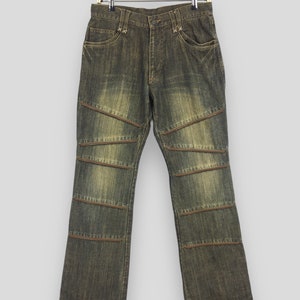 May include: Distressed denim jeans in a faded olive green hue, with distinctive horizontal brown stitching details across the knees and lower legs. The jeans are suspended from a gold-coloured hanger, showcasing their unique design.