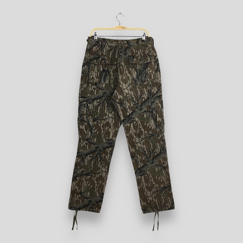 Pu&ograve; includere: Pantaloni cargo mimetici con un motivo boschivo nei toni del marrone, verde e grigio. I pantaloni sono appesi a una gruccia dorata. Hanno pi&ugrave; tasche e coulisse alle caviglie.
