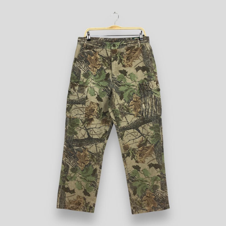 May include: Camouflage cargo pants with a button and zipper closure, featuring multiple pockets. The pants display a pattern of green, brown, and tan leaves and branches. The pants are hanging on a gold-colored hanger.