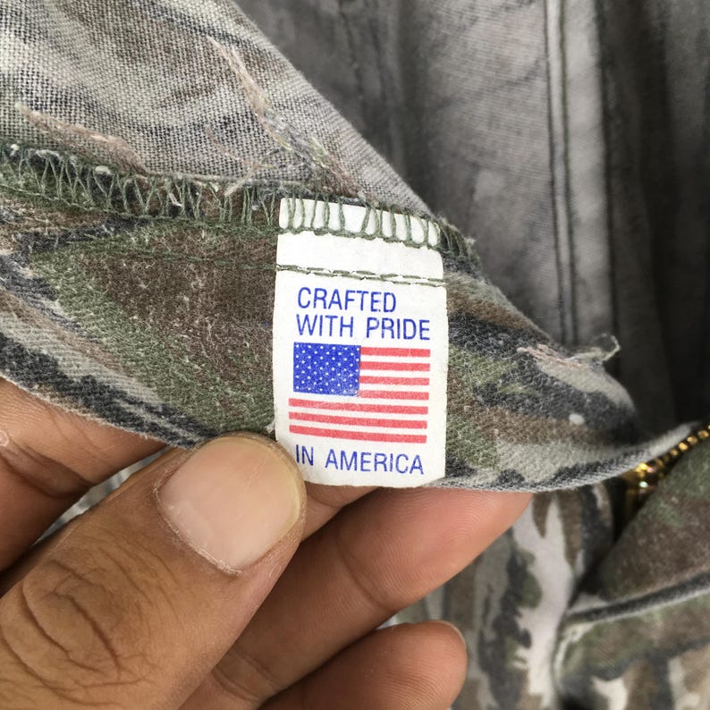 May include: Close-up of a tag with the text "CRAFTED WITH PRIDE" above an American flag graphic and the words "IN AMERICA" below. The tag is attached to camouflage fabric.