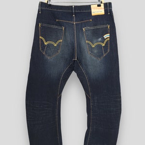May include: Dark wash denim jeans with gold stitching and unique pocket designs. The back pockets feature a distinctive wave-like pattern with gold stitching. A small tag reads "XV FUNCTION".