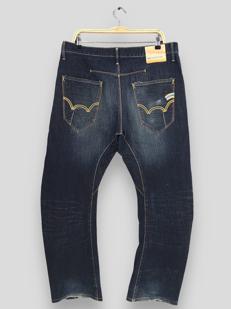 May include: Dark wash denim jeans with gold stitching and unique pocket designs. The back pockets feature a distinctive wave-like pattern with gold stitching. A small tag reads "XV FUNCTION".