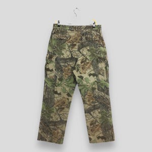 May include: Camouflage cargo pants with a realistic forest pattern in shades of green, brown, and tan. The pants have multiple pockets and are hanging on a hanger against a neutral background.