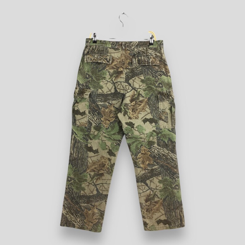 May include: Camouflage cargo pants with a realistic forest pattern in shades of green, brown, and tan. The pants have multiple pockets and are hanging on a hanger against a neutral background.