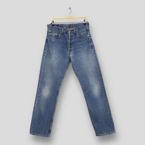 May include: Blue denim jeans hanging on a gold-colored hanger. The jeans have a button fly and a classic five-pocket design. The denim has a faded, worn appearance with lighter patches on the thighs and knees, suggesting wear. The jeans are a straight-leg style.