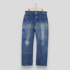 May include: Blue denim jeans with patches and distressed details hanging on a wooden hanger.