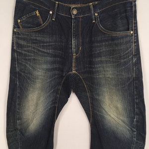 May include: Dark wash denim jeans with a faded effect. The jeans have a classic five-pocket design with a button and zipper closure. The stitching is a contrasting gold color, and the jeans are hanging on a gold hanger.