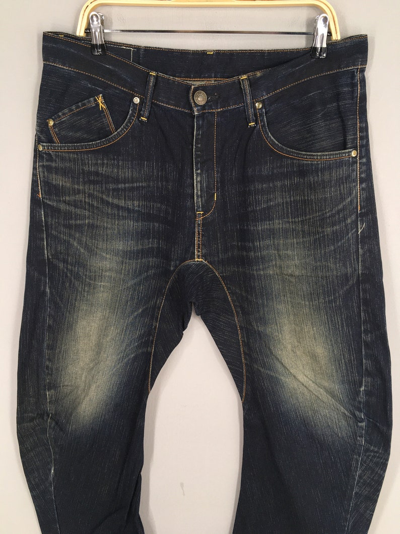 May include: Dark wash denim jeans with a faded effect. The jeans have a classic five-pocket design with a button and zipper closure. The stitching is a contrasting gold color, and the jeans are hanging on a gold hanger.