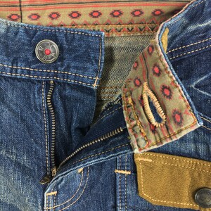 May include: Close-up of blue denim jeans with a partially unzipped zipper. The jeans feature decorative stitching, a button with a red center, and a patterned fabric detail on the waistband and pocket. The pocket has a brown suede patch.