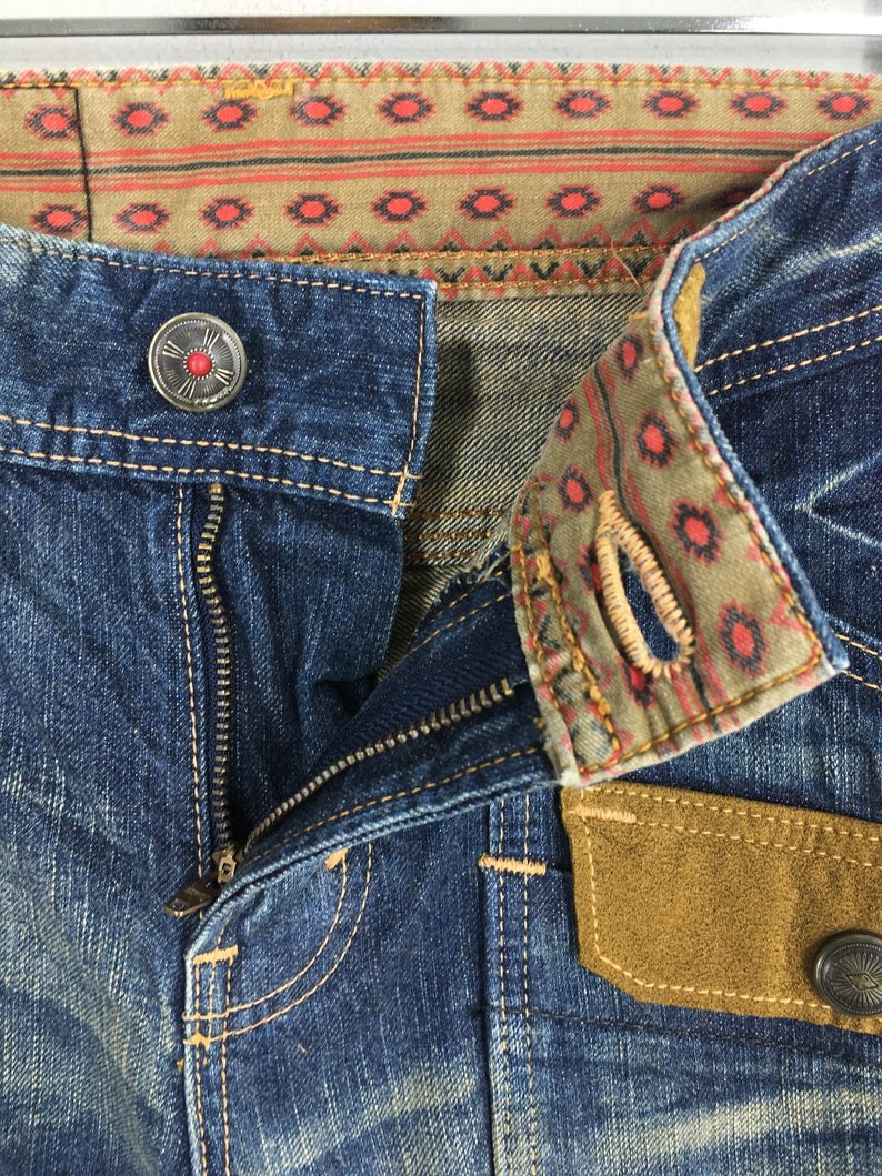 May include: Close-up of blue denim jeans with a partially unzipped zipper. The jeans feature decorative stitching, a button with a red center, and a patterned fabric detail on the waistband and pocket. The pocket has a brown suede patch.