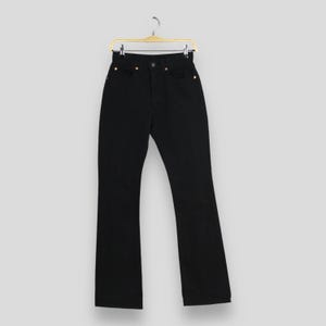 May include: A pair of solid black high-waisted denim jeans hanging on a gold-toned metal hanger. The jeans feature a classic five-pocket design with visible copper-coloured rivets and a central button closure at the waist, showcasing a straight-leg cut.