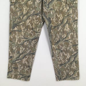 May include: Camouflage cargo pants in shades of brown, tan, and green. The pants feature a detailed tree branch pattern and include cargo pockets. The fabric appears to be a durable material, suitable for outdoor activities.