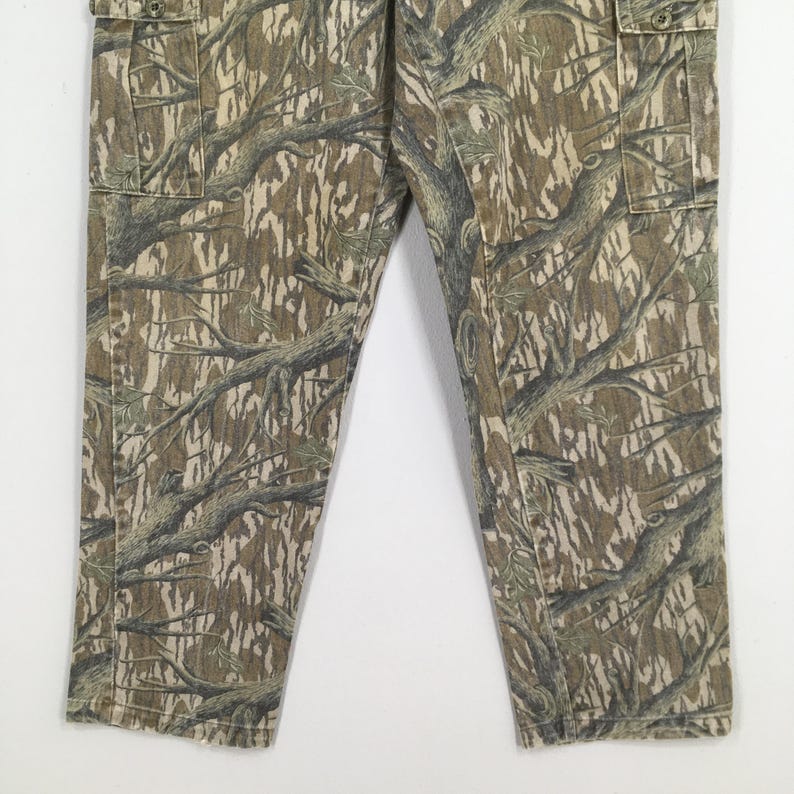 May include: Camouflage cargo pants in shades of brown, tan, and green. The pants feature a detailed tree branch pattern and include cargo pockets. The fabric appears to be a durable material, suitable for outdoor activities.
