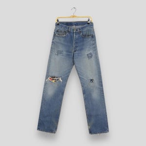May include: Light blue denim jeans with distressed details, including patches and a large rip on the left knee. The jeans are hanging on a gold-colored hanger. The jeans have a button fly and classic five-pocket design.