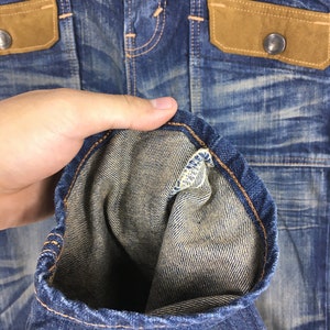 May include: Close-up of a blue denim jacket with brown suede-like pocket flaps and silver button closures. The jacket has a worn, distressed appearance with visible stitching and a folded sleeve, showcasing the interior fabric.