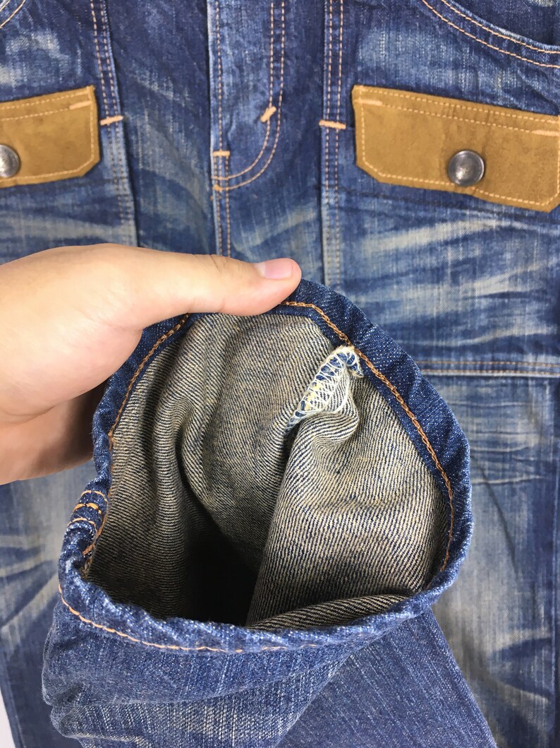 May include: Close-up of a blue denim jacket with brown suede-like pocket flaps and silver button closures. The jacket has a worn, distressed appearance with visible stitching and a folded sleeve, showcasing the interior fabric.