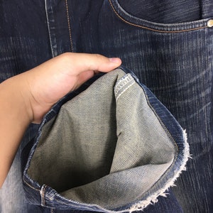 May include: Close-up of dark blue denim jeans. The jeans have a classic five-pocket design with a button and zipper closure. The inside of the jeans is a lighter shade of blue, and the hem is frayed. The jeans are held open to show the inside.