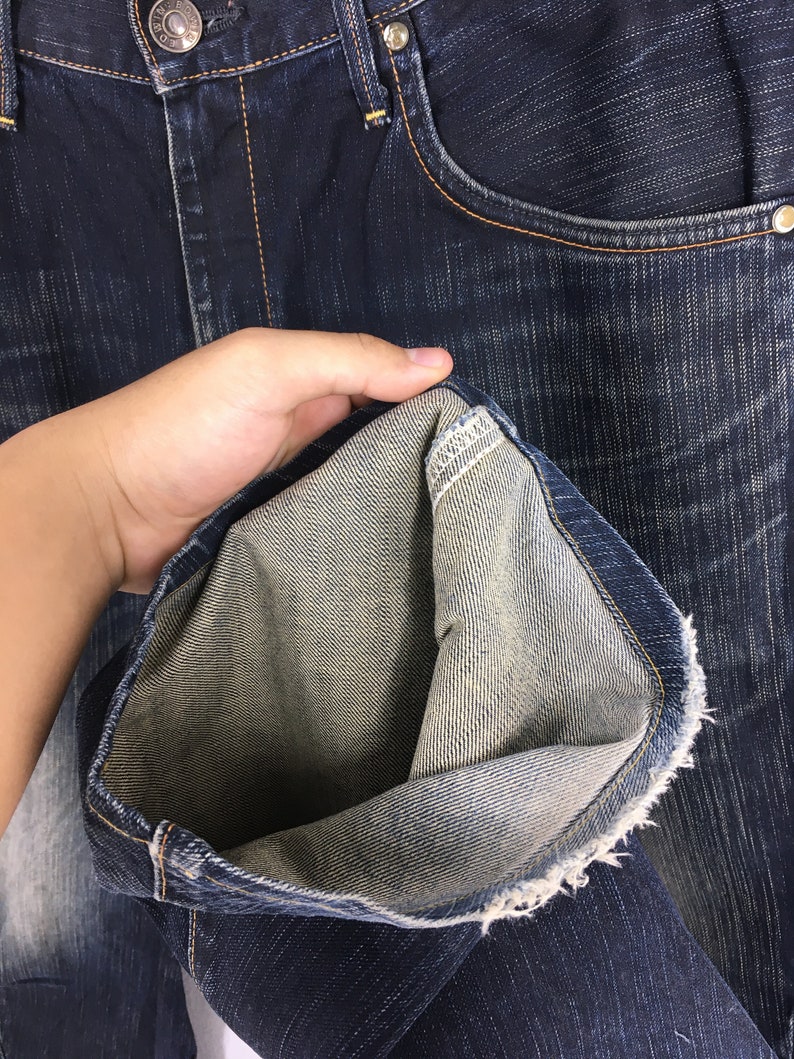 May include: Close-up of dark blue denim jeans. The jeans have a classic five-pocket design with a button and zipper closure. The inside of the jeans is a lighter shade of blue, and the hem is frayed. The jeans are held open to show the inside.
