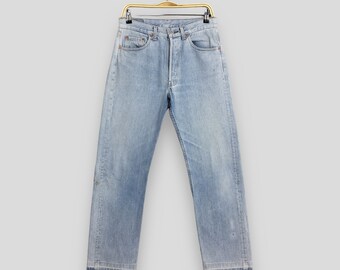 May include: Light blue denim jeans hanging on a gold-colored hanger. The jeans have a button fly, classic five-pocket design, and a straight leg. Faded areas and distressed details add character to the vintage style.