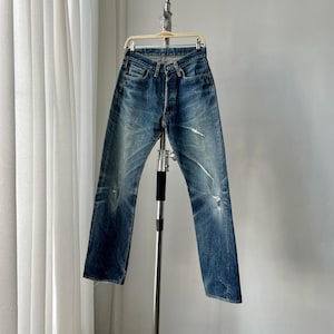 May include: Faded blue denim jeans with a button fly and distressed details. The jeans are hanging on a metal hanger, showcasing a classic, worn-in look. The denim has light fading and some rips.