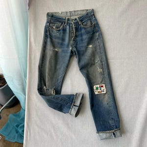 Könnte beinhalten: Blaue Jeans aus Denim mit Knopfleiste, abgenutzten Stellen und einem kleinen rechteckigen Aufnäher am linken Bein mit einer bunten Stickerei aus abstrakten Formen in Rot, Grün und Blau. Die Beinabschlüsse sind umgekrempelt und zeigen die Naht.