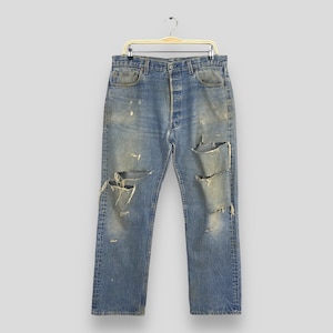 May include: Distressed blue denim jeans with multiple rips and tears throughout the fabric. The jeans are hanging on a wooden hanger.
