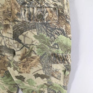 May include: Close-up of camouflage pants with a buttoned pocket. The pants feature a pattern of green, brown, and tan foliage, creating a realistic woodland design. The fabric appears to be a durable material, suitable for outdoor activities.