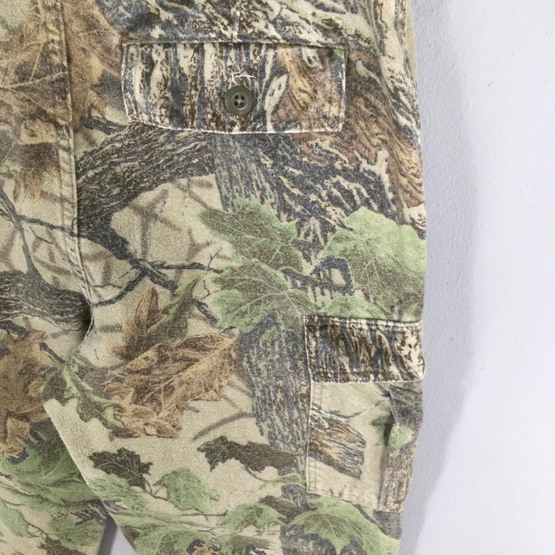 May include: Close-up of camouflage pants with a buttoned pocket. The pants feature a pattern of green, brown, and tan foliage, creating a realistic woodland design. The fabric appears to be a durable material, suitable for outdoor activities.