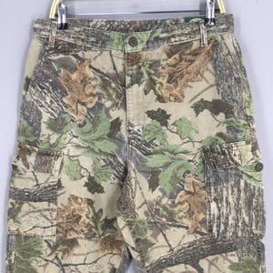 May include: Camouflage cargo pants with a button closure and multiple pockets. The pants feature a pattern of green, brown, and tan leaves and branches. The pants are hanging on a metal hanger.
