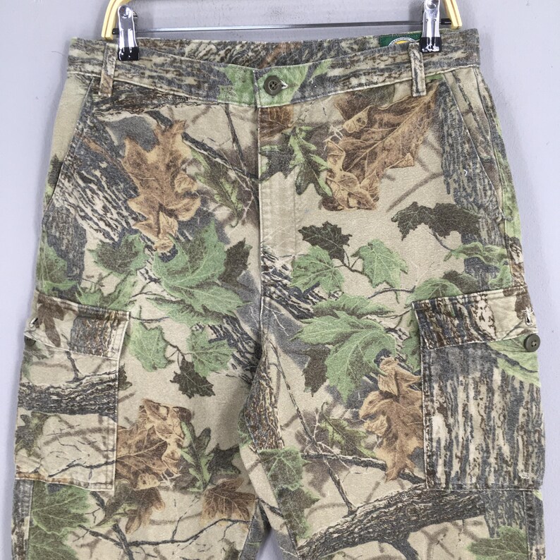 May include: Camouflage cargo pants with a button closure and multiple pockets. The pants feature a pattern of green, brown, and tan leaves and branches. The pants are hanging on a metal hanger.