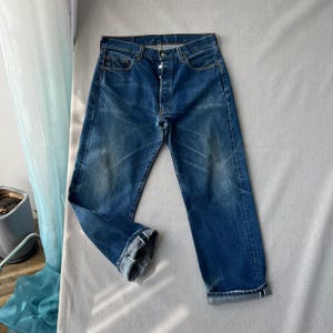 Könnte beinhalten: Eine dunkelblaue Jeans aus Denim mit Knopfleiste, fünf Taschen und geradem Bein. Der Stoff weist leichte Verwaschungen und Sitzfalten auf, wobei beide Hosenbeine am Saum umgeschlagen sind, um die innenliegende Webkante sichtbar zu machen.