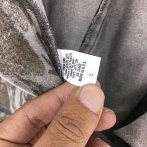 May include: Close-up of a gray and brown camouflage-patterned shirt. The shirt's tag reads "ITEM-0004, TURN INSIDE OUT, WASH COLD WATER, HANG TO DRY, 100% COTTON, RN 73342, MADE IN U.S.A., L".