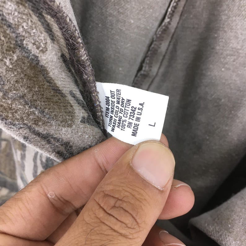 May include: Close-up of a gray and brown camouflage-patterned shirt. The shirt's tag reads "ITEM-0004, TURN INSIDE OUT, WASH COLD WATER, HANG TO DRY, 100% COTTON, RN 73342, MADE IN U.S.A., L".