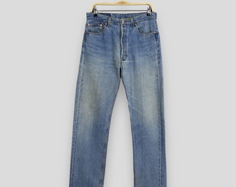 May include: Light blue denim jeans hanging on a gold-colored hanger. The jeans have a button fly and classic five-pocket design. The denim has a faded, worn appearance, with subtle variations in color.