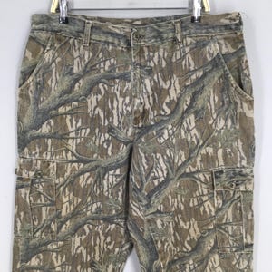 May include: Camouflage cargo pants with a button closure and multiple pockets. The pants feature a brown, green, and tan tree branch pattern. The pants are hanging on a metal hanger.