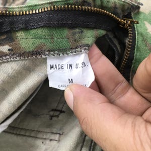Puede incluir: Primer plano de pantalones de camuflaje con cremallera visible y etiqueta que dice "MADE IN U.S.A.". La etiqueta tambi&eacute;n muestra la talla "M". La tela tiene un patr&oacute;n verde, marr&oacute;n y beige.