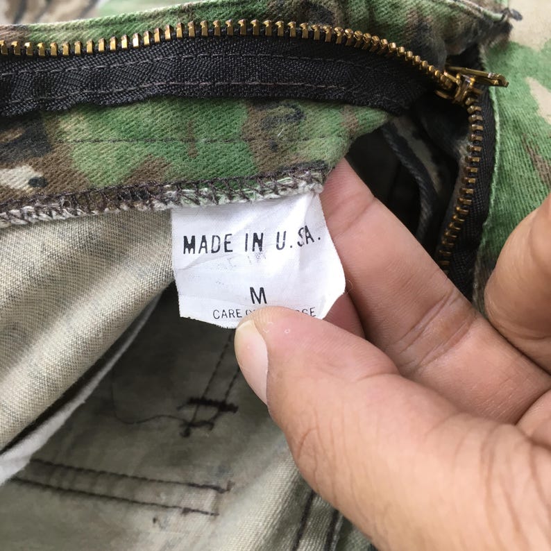 Puede incluir: Primer plano de pantalones de camuflaje con cremallera visible y etiqueta que dice "MADE IN U.S.A.". La etiqueta tambi&eacute;n muestra la talla "M". La tela tiene un patr&oacute;n verde, marr&oacute;n y beige.